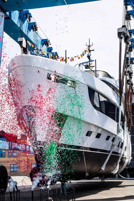 Azimut Grande 44M super yatının suya indirme töreninde, devasa beyaz ve siyah gövdeli teknenin etrafına kırmızı ve yeşil konfetiler saçılırken, mavi bir gantry vinci tarafından nazikçe havada asılı tutulduğu ve altından dumanlar çıktığı görünüyor.