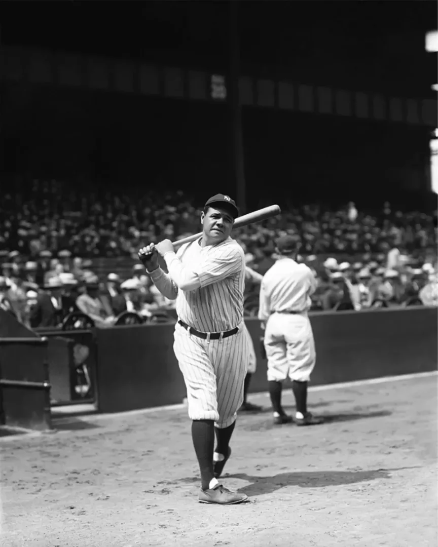 Babe Ruth, beysbol sahasında üzerinde klasik çizgili forması ve beyzbol sopasıyla poz verirken, arka planda seyircilerin olduğu stadyumun tribünleri görünen bu siyah beyaz fotoğrafta efsanevi kariyerinin bir anını yansıtıyor.