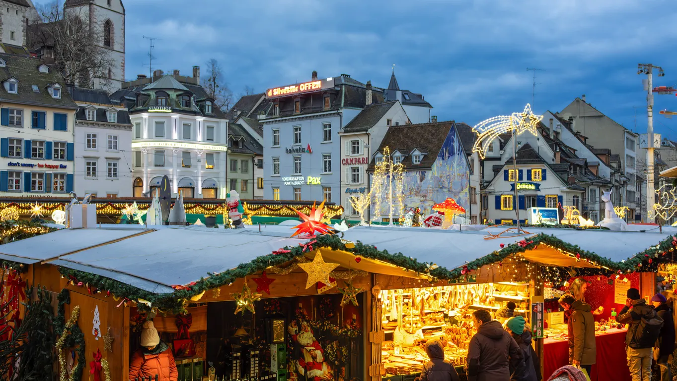 Basel'deki bir Noel pazarında, karla kaplı çatılara sahip, ışıklı çelenkler ve yıldızlarla süslenmiş ahşap tezgahların arasında dolaşan insanların oluşturduğu sıcak ve şenlikli atmosfer, arka plandaki tarihi binalar ve göz alıcı ışıklandırmalarla tamamlanıyor.