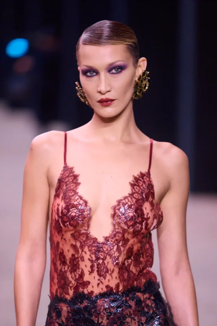 Bella Hadid, Saint Laurent podyumunda mor tonlarında iddialı göz makyajı ve bordo rujuyla, ıslak görünümlü geriye taranmış saçları ve gösterişli altın rengi küpeleriyle zarif bir şekilde yürürken, derin yakalı kırmızı dantel bluzuyla dikkat çekiyor.