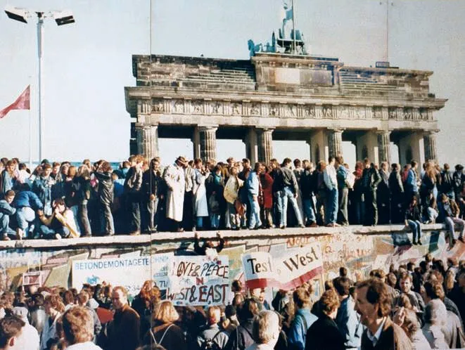1989 yılında Berlin Duvarı'nın yıkılışını kutlayan coşkulu kalabalıklar, üzerinde grafiti ve OVER PEACE IN WEST & EAST gibi mesajlar bulunan duvarın üstünde ve önünde, tarihi Brandenburg Kapısı'nın görkemli silüeti altında bir araya gelmiş.