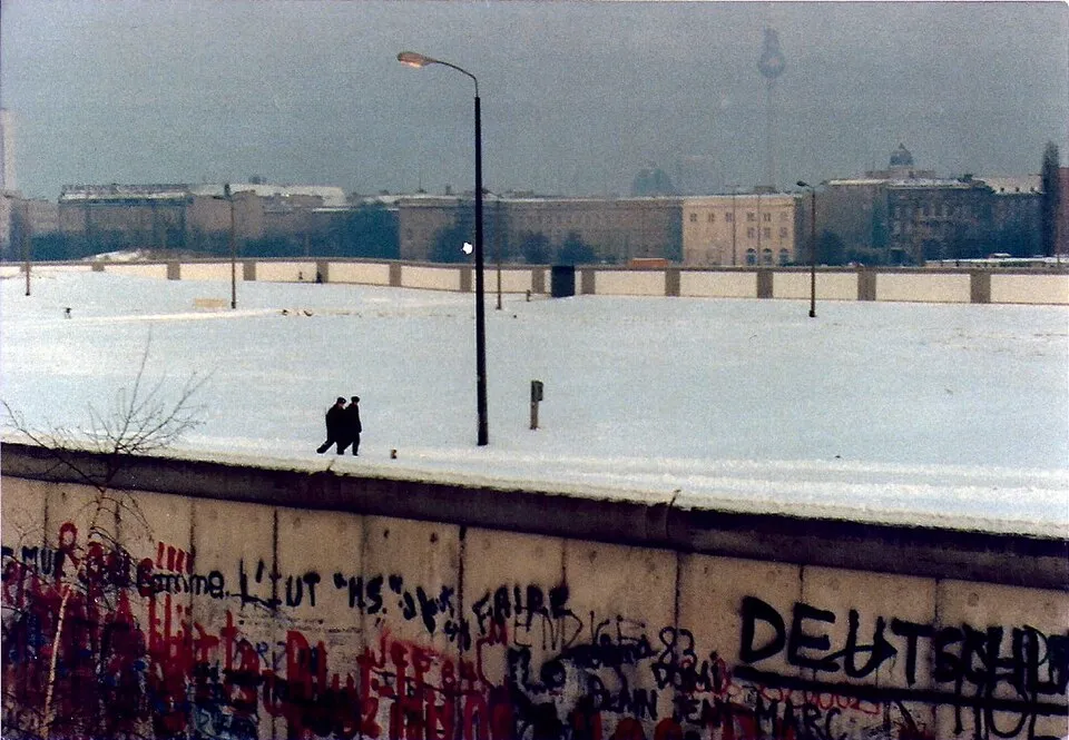 Berlin Duvarı'nın rengarenk grafitiyle kaplı beton bariyerinin ön planda olduğu, karla kaplı geniş alanda yürüyen iki kişinin siluetinin ve uzaklarda puslu Berlin şehir manzarasının göründüğü bu görüntü, duvarın ilginç bilgilerini düşündürüyor.