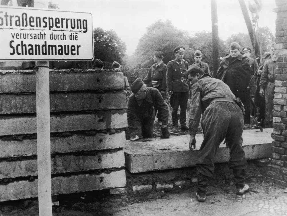 Almanca Strassensperrung verursacht durch die Schandmauer (Utanç Duvarı nedeniyle yol kapalı) yazan bir tabelanın önünde, işçilerin beton blokları üst üste dizerek bir bariyer oluşturduğu ve çevrede üniformalı askerlerin denetim yaptığı bu siyah beyaz kare, Berlin Duvarı'nın yapım anlarını ve o dönemin gergin atmosferini yansıtıyor.