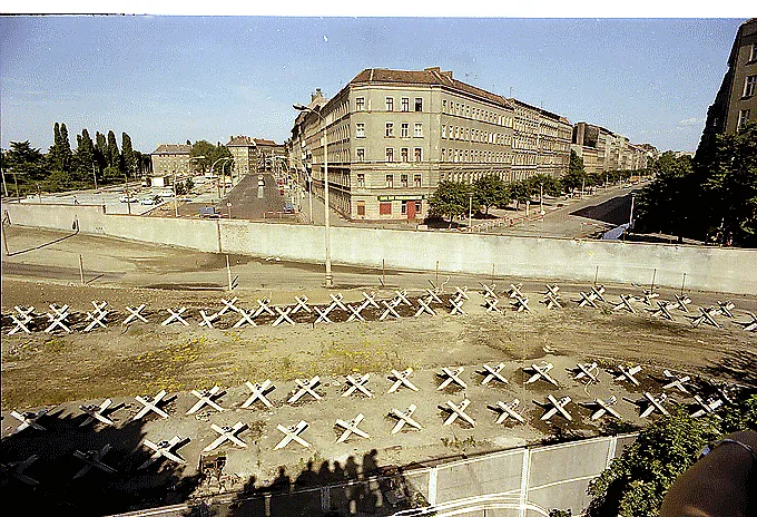 Kuşbakışı bir açıyla çekilmiş bu tarihi fotoğrafta, Berlin Duvarı'nın önündeki geniş boş toprak alanda sıralanmış çok sayıda X şeklinde tank savar engel, duvarın ardında ise binalar ve araçların olduğu bir cadde uzanıyor.