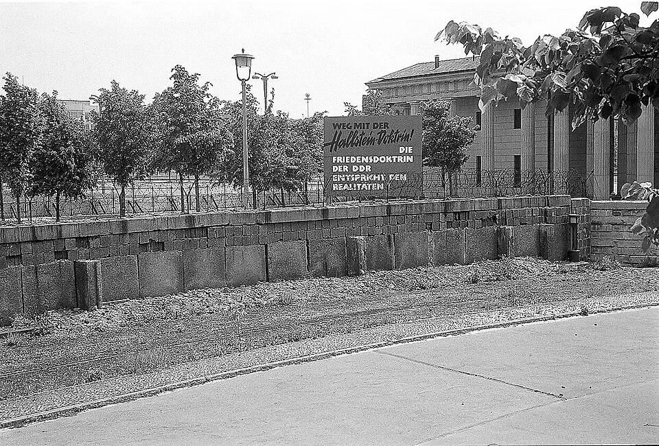 Siyah beyaz tarihi bir fotoğrafta, Hallstein Doktrini'ni hedef alan Almanca bir pankartın önünde yükselen, üstünde dikenli teller bulunan beton Berlin Duvarı parçası, ağaçlar ve sütunlu bir bina belirginleşerek, soğuk savaş döneminin ayrılık ve gerilimini yansıtıyor.