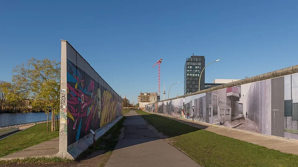 Berlin Duvarı'nın Spree Nehri kıyısında, bir tarafı renkli grafiti sanatı, diğer tarafı ise duvarla ilgili eski fotoğraflarla kaplı uzun bir bölümünün, arka planda Berlin Fernsehturm'u ve modern binaların yükseldiği, güneşli bir günde çekilmiş, ortasında bir yürüyüş yolunun bulunduğu geniş açıdan görünümü.