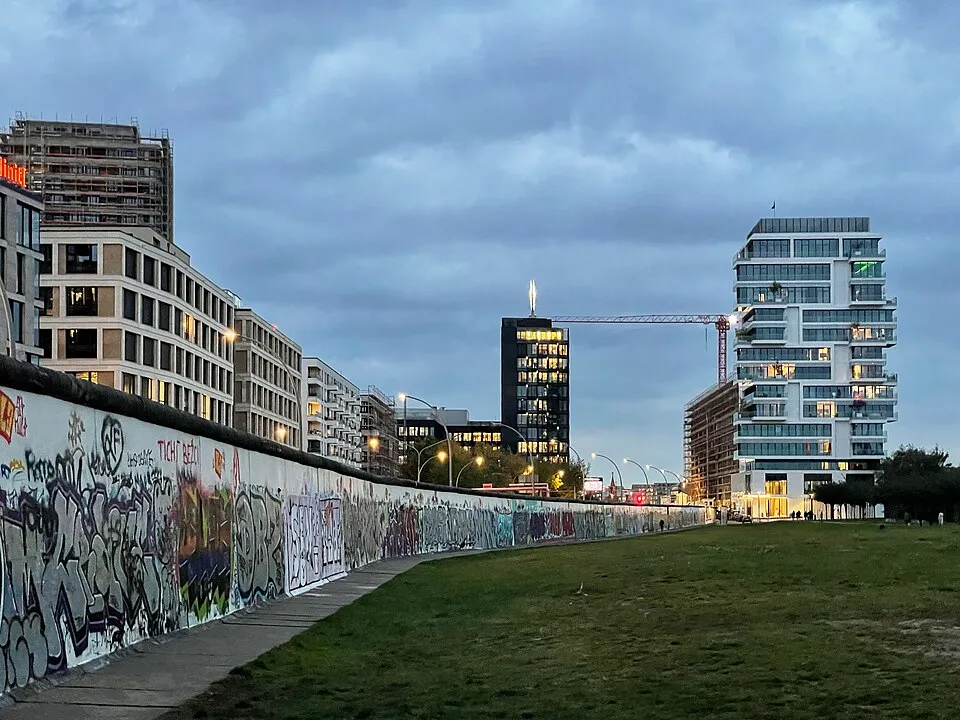 Grafiti kaplı Berlin Duvarı, önünde uzanan yeşil bir çimenlik alan ve ardında yükselen modern binalar, inşaat halindeki yapılar ile onları birbirine bağlayan kırmızı bir vinçle, şimdiki halini yansıtan bulutlu bir havada görülüyor.