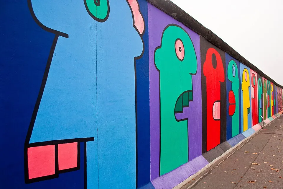 Berlin Duvarı üzerinde, Thierry Noir'ın kendine özgü tarzıyla mavi, yeşil, mor ve kırmızı tonlarda sıralanmış soyut insan figürlerini gösteren uzun bir duvar resmi, yanında düşmüş yapraklarla dolu bir yol ile gri gökyüzü altında uzanıyor.