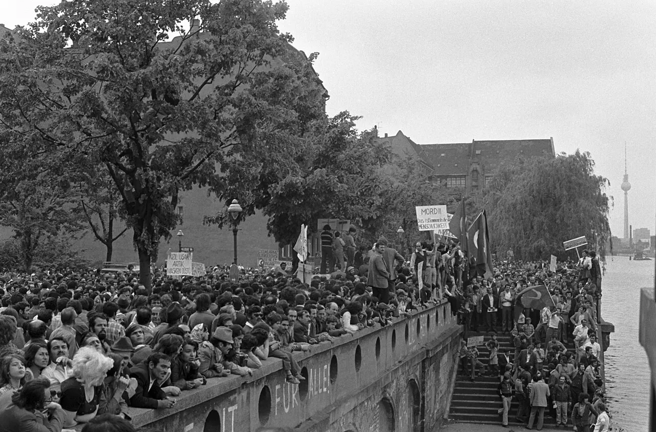 Berlin Duvarı dönemiyle ilişkilendirilebilecek bir protestoda, köprü kenarında toplanmış, Türk bayrakları ve üzerinde Kızıl Uşak artık masken düştü yazan pankartlar taşıyan kalabalık bir Türk grubu, arka planda Berlin TV Kulesi'nin siluetini gösteren siyah beyaz bir fotoğraf.