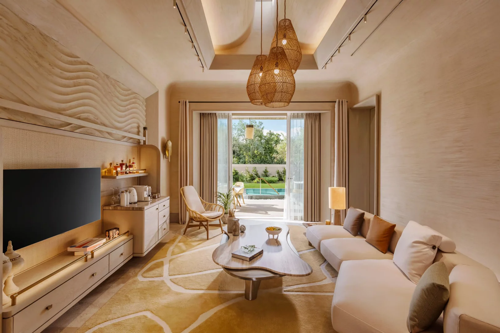 BIJAL villalarının konforlu ve geniş yaşam alanını gözler önüne seren bu karede, modern bir kanepe, özgün tasarımlı ahşap sehpa, şık bir minibar ünitesi ve dalgalı dokulu duvar detayı dikkat çekerken, büyük cam kapılar dışarıdaki yemyeşil bahçe ve özel havuz ile bütünleşerek Akdeniz'in dingin atmosferini içeri taşıyor.