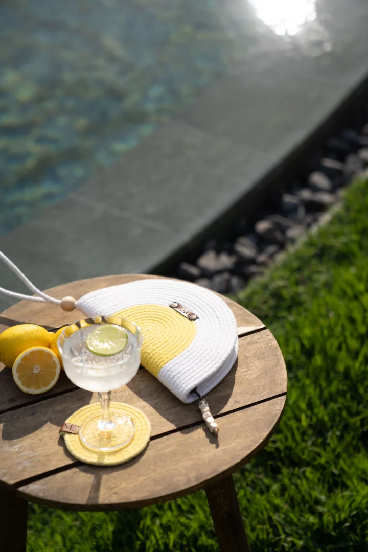 BIJAL'in lüks villa atmosferinde, özel havuz başında konumlanan ahşap bir sehpada, serinletici bir içecek, limonlar ve sarı beyaz bir çanta güneşli bir Akdeniz gününün keyfini çıkarmanın kişiselleştirilmiş anlarını yansıtıyor.
