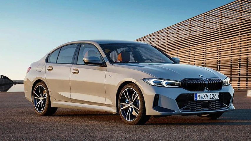 Modern ahşap panelli mimariye sahip bir yapının ve uzaklaşan köprülü su manzarasının önünde, şık metalik bej gri rengindeki yeni nesil BMW 3 Serisi otomobil, karartılmış jantları ve keskin çizgileriyle dikkat çekici bir şekilde park etmiş görünüyor.