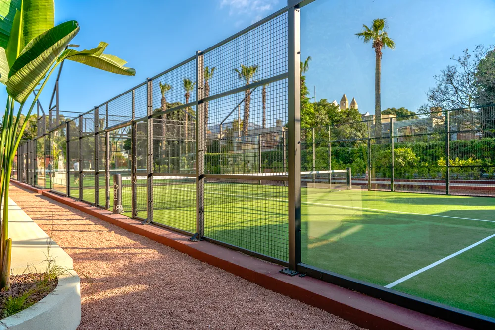 Bodrum'un mesh çitler ve şeffaf cam panellerle çevrili, parlak yeşil suni çim zeminli padel kortlarından birini gösteren bu geniş açılı görüntüde, kortun solunda çakıllı bir yürüyüş yolu ve tropikal bitkiler, arka planda ise palmiye ağaçları ile eski bir bina gökyüzüne uzanıyor.