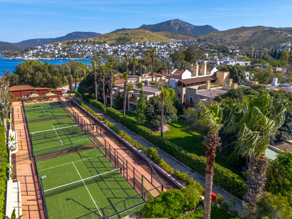 Bodrum'da palmiye ağaçları ve yemyeşil bir çevrede konumlanmış, masmavi denizin ve uzaklardaki dağların eşlik ettiği, canlı yeşil renkli birden fazla padel kortu güneşli bir havada yukarıdan görünüyor.