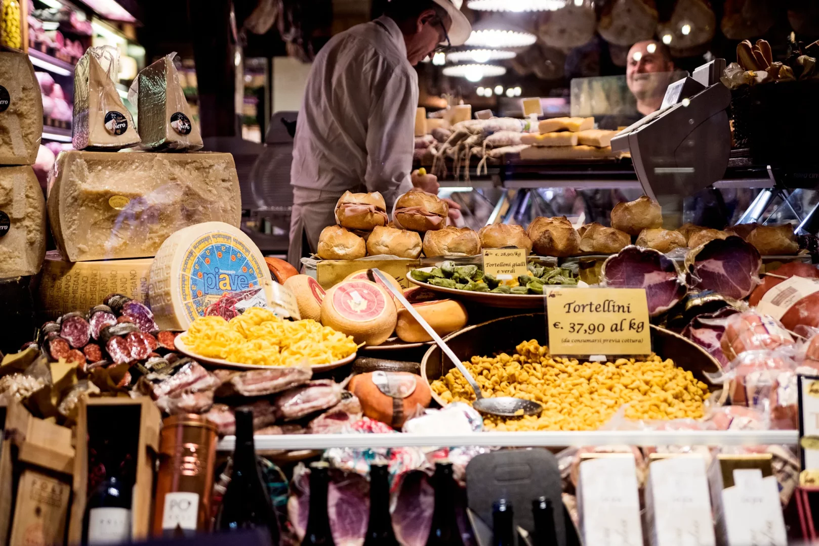Bologna'nın hareketli bir gıda pazarında, peynir tekerlekleri, dilimlenmiş şarküteri ürünleri, taze tortellini çeşitleri ve ekmek arası sandviçler gibi çeşitli geleneksel İtalyan lezzetleriyle dolu tezgahın arkasında bir çalışan ürünleri düzenliyor.