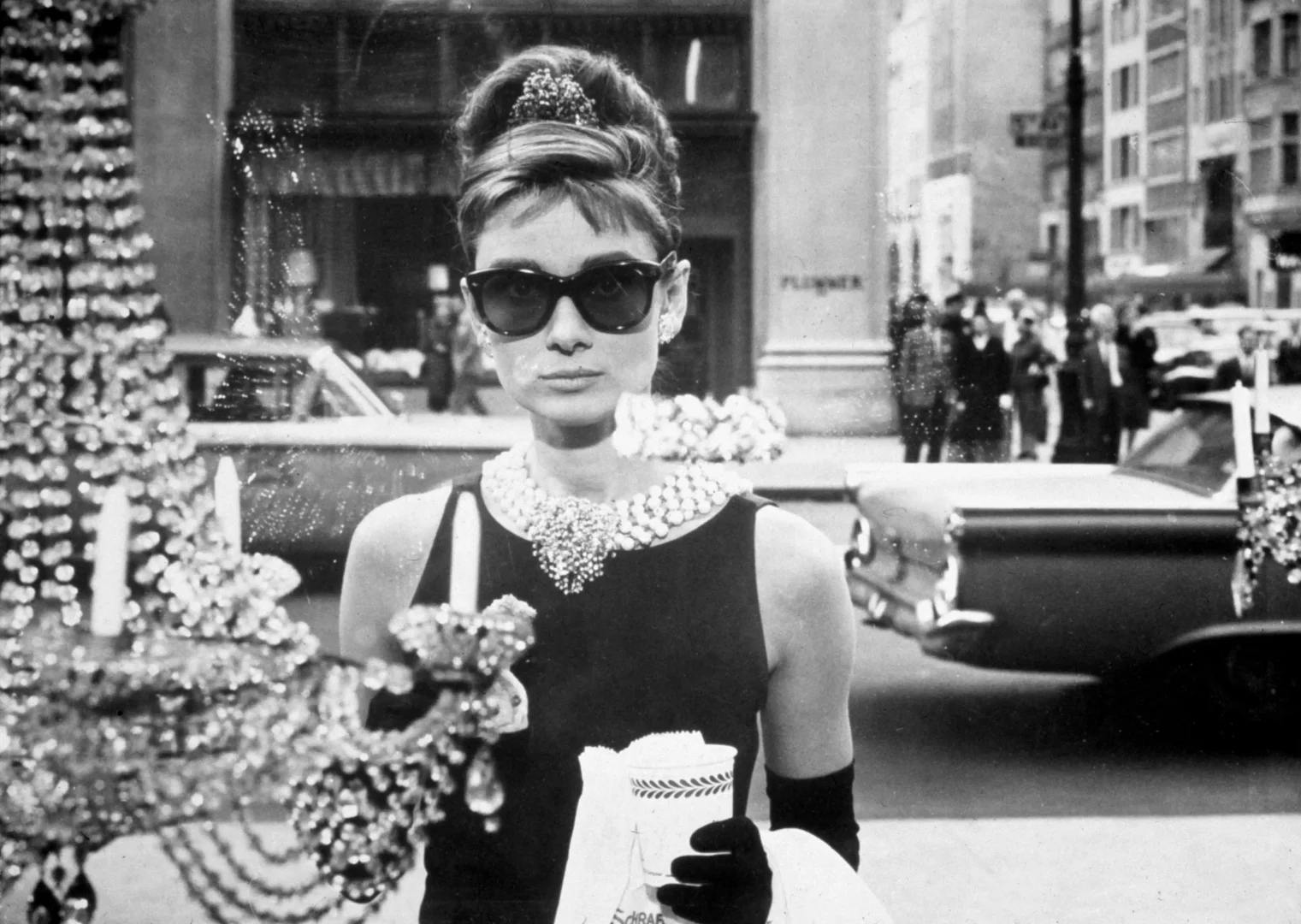 Siyah beyaz bir karede, güneş gözlüklü, topuz saçlı ve gösterişli inci-taş kolyesiyle Audrey Hepburn, Tiffany'de Kahvaltı filmindeki Holly Golightly karakteri olarak elinde kahve ve kruvasanla, New York'ta bir Tiffany & Co. mağazasının vitrini önünde durarak, ikonik stilini ve markanın zarafetini yansıtıyor.
