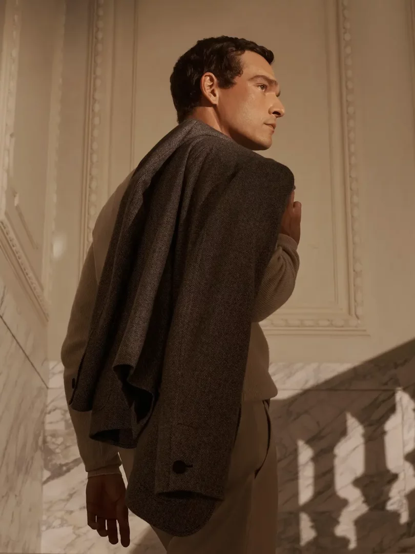 Brioni blazer kampanyasından zarif bir görselde, hafif bej bir kazak ve pantolon giymiş, omzunda şık, koyu gri balıksırtı desenli bir blazer taşıyan, sağa doğru profilden bakan ve klasik mermer detaylı bir odada duran şık bir adam.