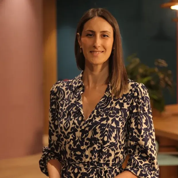 Büşra Tarcalır, uzun kahverengi saçları, sıcak gülümsemesi ve lacivert ile krem rengi damask desenli şık elbisesiyle kameraya dönük poz verirken, pembe ve lacivert tonlarının hafifçe bulanıklaştığı bir arka plan önünde zarif bir portre.
