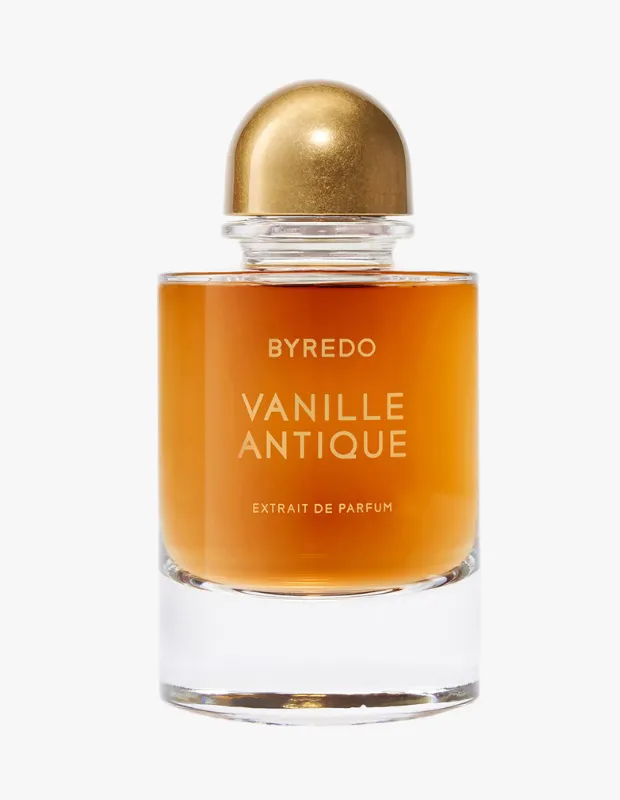 Parlak altın rengi kubbeli kapağı ve kalın şeffaf cam tabanı olan, içinde sıcak kehribar rengi Byredo Vanille Antique extrait de parfum sıvısı bulunan bir parfüm şişesi, beyaz bir arka plan önünde duruyor.