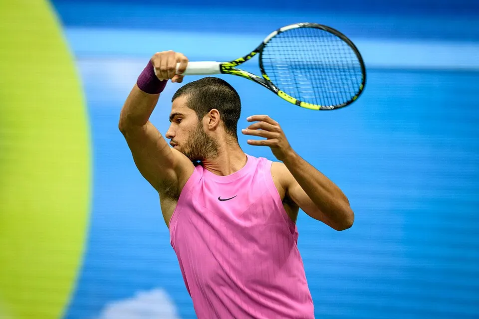 Carlos Alcaraz, pembe spor tişörtü ve sol bileğindeki mor bantla, başının üzerinden tenis raketini çevirerek topa vurmaya odaklanmış, dinamik bir karede görünüyor. Arka plandaki bulanık mavi zemin ve yeşil detaylar, Amerika Açık gibi büyük bir turnuvadaki yoğun maç anını vurguluyor.
