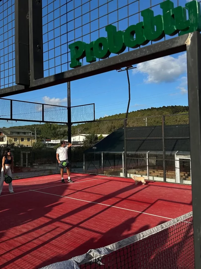 Casa Kilyos İstanbul padel kortlarından birinde, kırmızı zeminli sahada raketleriyle duran iki oyuncu ve yanlarında dolaşan sevimli bir köpek, güneş ışınlarının ağlara düşen gölgeleriyle aydınlanan çitli bir alanda keyifli bir an yaşarken, çitlerin üst kısmında yeşil Prob yazısı ve arka planda binalar ile yemyeşil ağaçlar görünüyor.