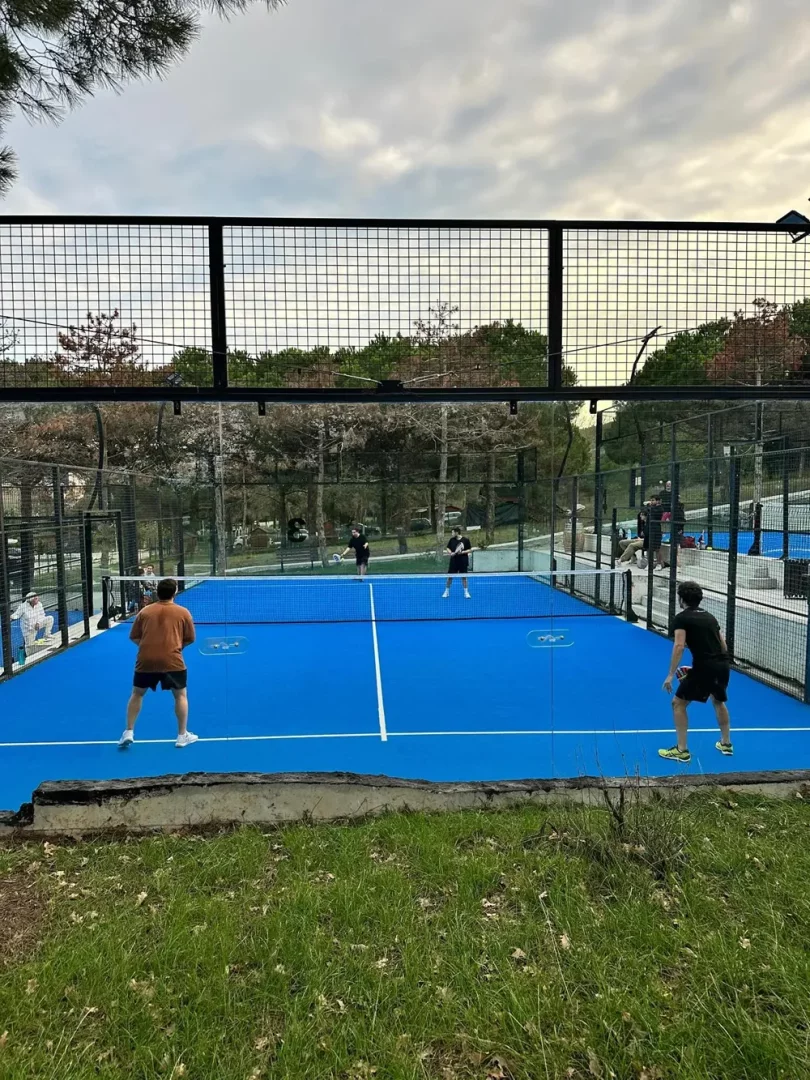 Mavi zeminli, etrafı tel örgülerle çevrili açık havada bir padel kortunda dört oyuncu dinamik bir şekilde padel oynarken görülüyor; arka planda ağaçlar ve bulutlu gökyüzü, bu modern tesisin Kilyos, İstanbul gibi doğal bir ortamda konumlandığını düşündürüyor.