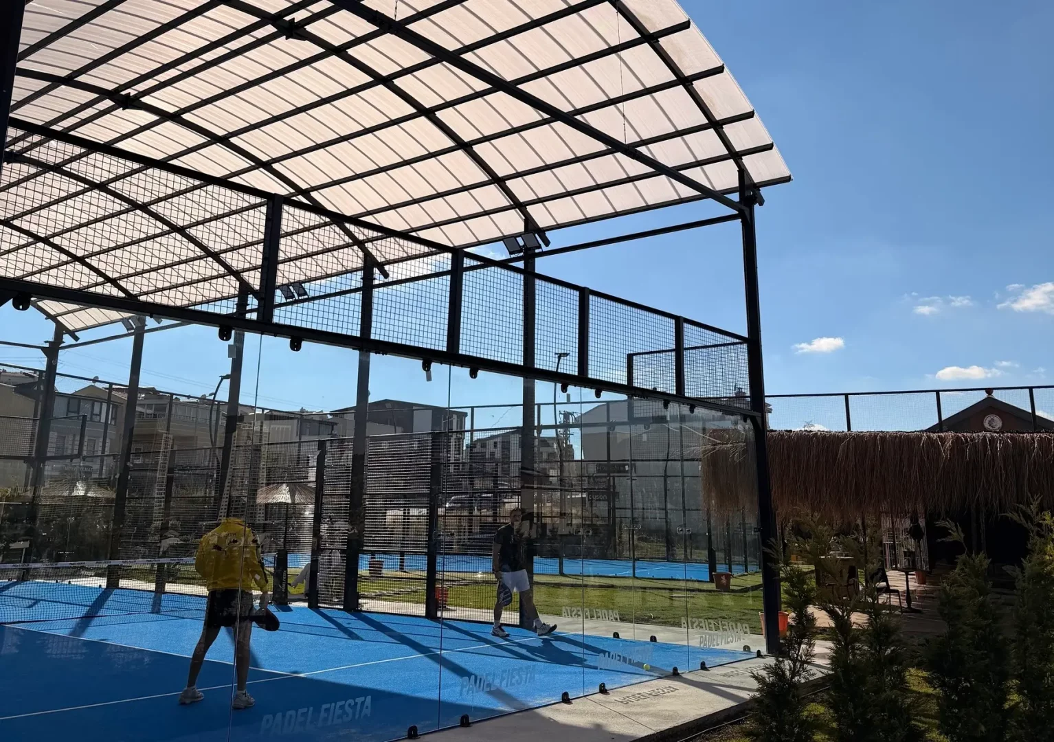 Güneşli bir günde, şeffaf cam duvarlar ve metal kafeslerle çevrili mavi yüzeyli Padel Fiesta çatılı kortunda iki kişi padel oynarken görülüyor; kortun üzeri beyaz yarı şeffaf bir çatı ile kapatılmış ve çevrede başka kortlar ile yeşil alanlar mevcut.
