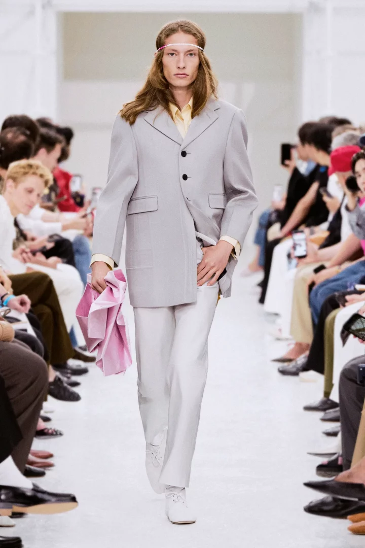 Celine İlkbahar 2027 erkek giyim defilesinde, uzun dalgalı saçları ve başında ince pembe bandı olan bir model, açık gri oversized bir blazer, sarı gömlek ve beyaz pantolon kombinasyonuyla podyumda yürürken sol elinde pembe bir kumaş parçası taşıyor.