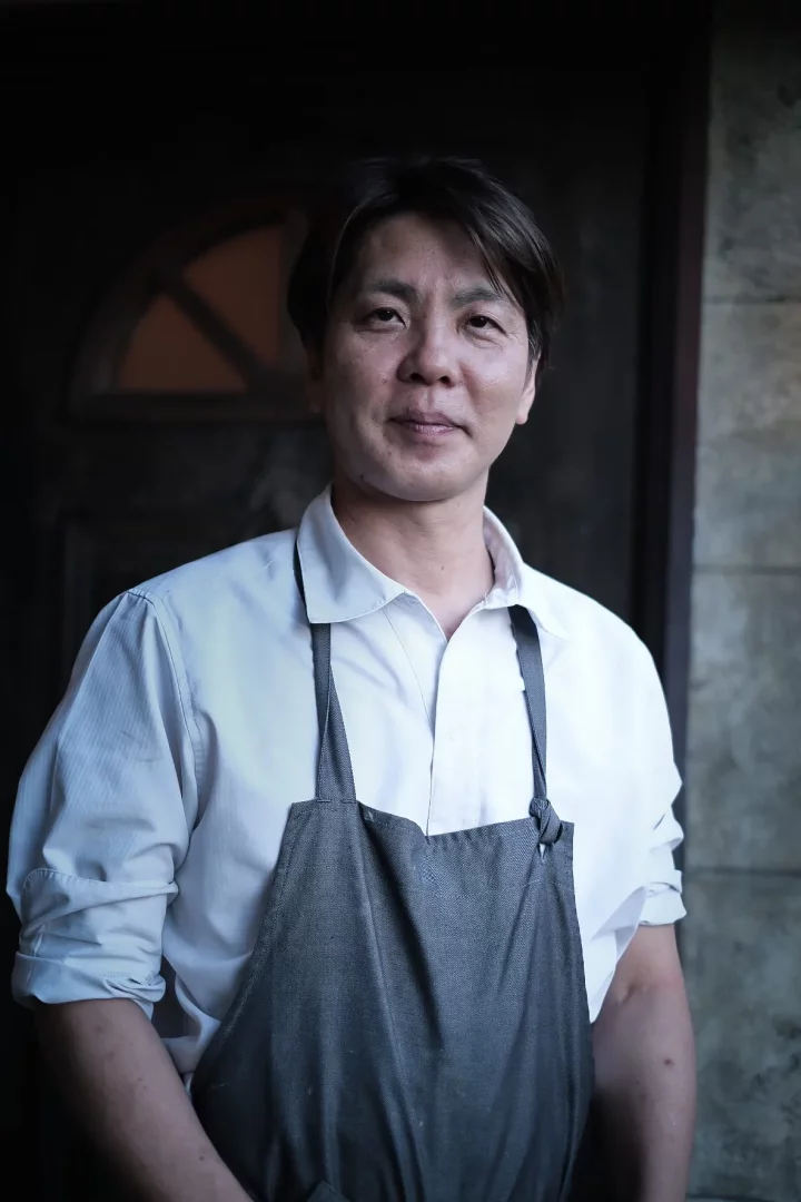 Michelin yıldızlı şef Kanji Kobayashi, kolları sıvalı beyaz gömleği ve gri önlüğüyle, karanlık arka plan önünde hafifçe gülümseyerek kameraya bakarken, İstanbul'a özel tadım menüsü ve Agrigastronomy yaklaşımını temsil ediyor.