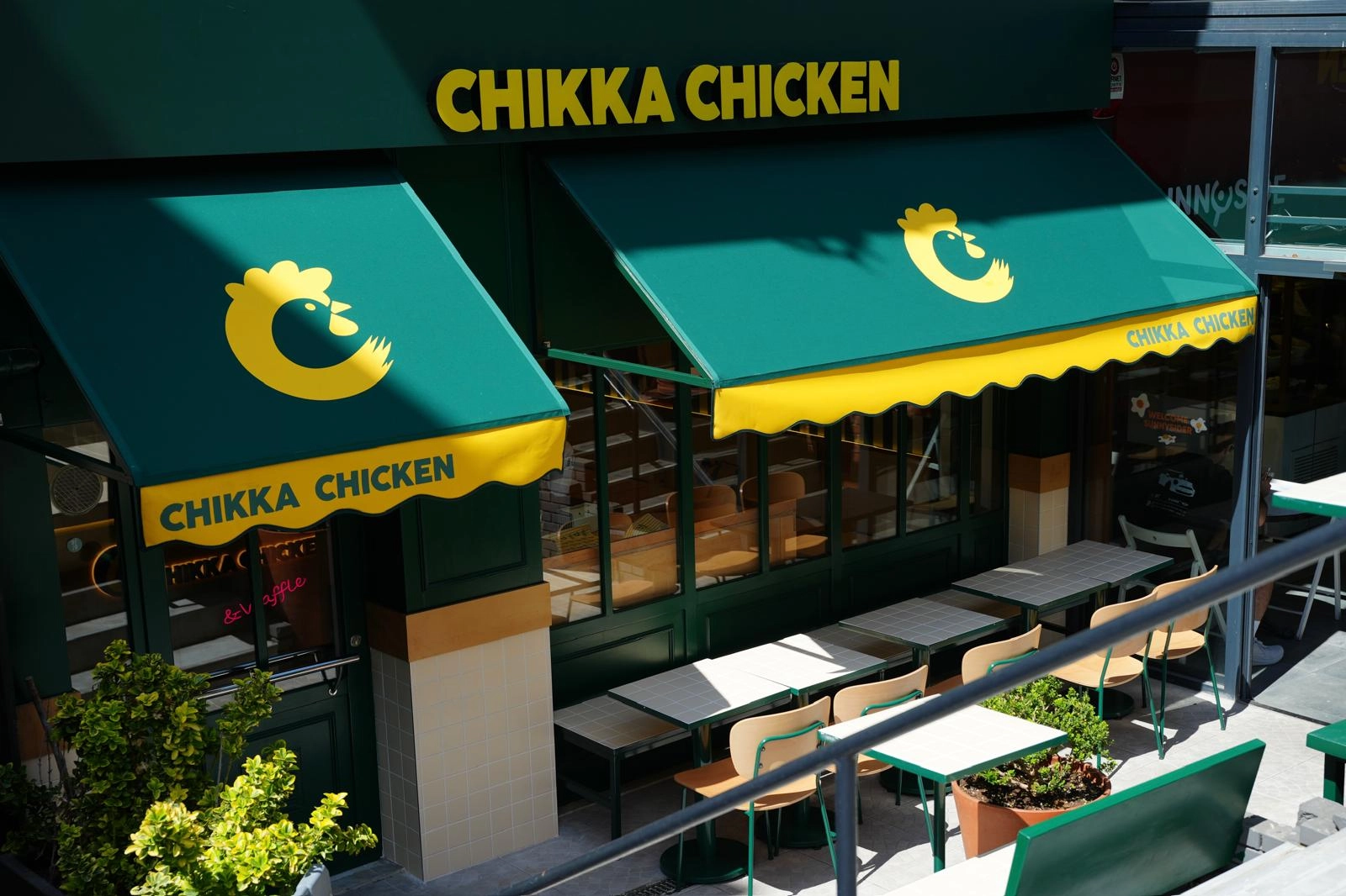 Chikka Chicken restoranının koyu yeşil ve parlak sarı renklerde tasarlanmış dış cephesi, üstünde marka adı ve logosu olan tenteleri, ayrıca beyaz masa ve ahşap sandalyeleriyle düzenlenmiş davetkar dış oturma alanını gösteriyor.
