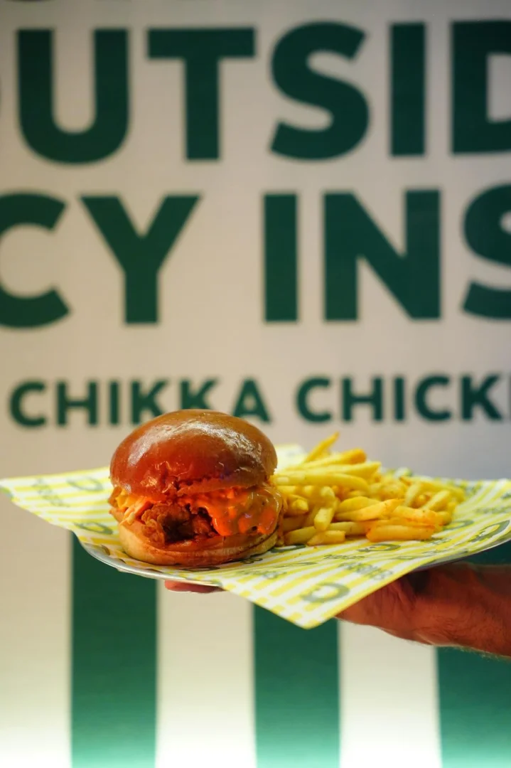 Bir el, parlak sarı desenli bir tabakta, baharatlı turuncu sosla cömertçe gezdirilmiş parlak ekmekli bir Chikka Chicken burgerini ve yanında altın rengi patates kızartmalarını tutarken, arka planda yeşil harflerle bulanıklaşmış OUTSIDE SPICY INSIDE CHIKKA CHICKEN yazısı dikkat çekiyor.