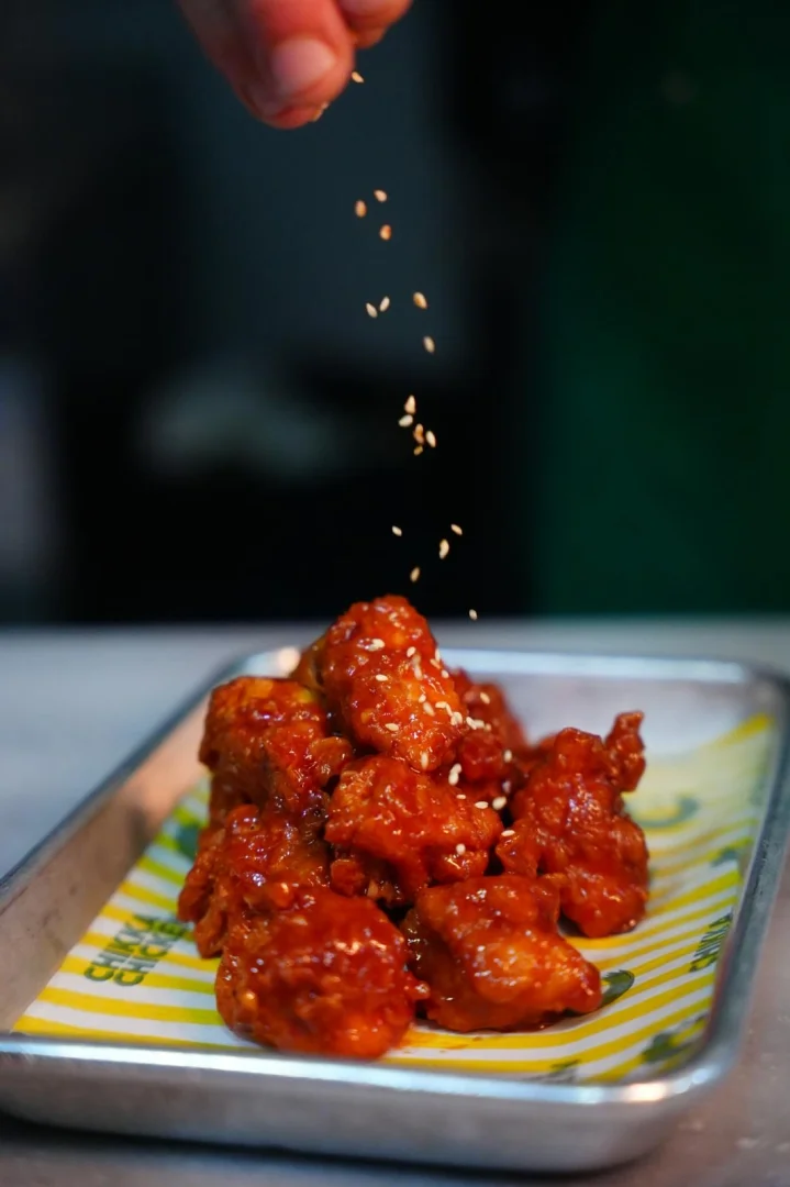 Metal bir tepsideki sarı çizgili kağıt üzerinde duran, parlak ve kırmızı sosla kaplı tavuk parçalarının üzerine bir elin susam serptiği bu görüntü, Chickka Chicken'ın enfes soslu tavuklarını iştah açıcı bir şekilde sergiliyor.