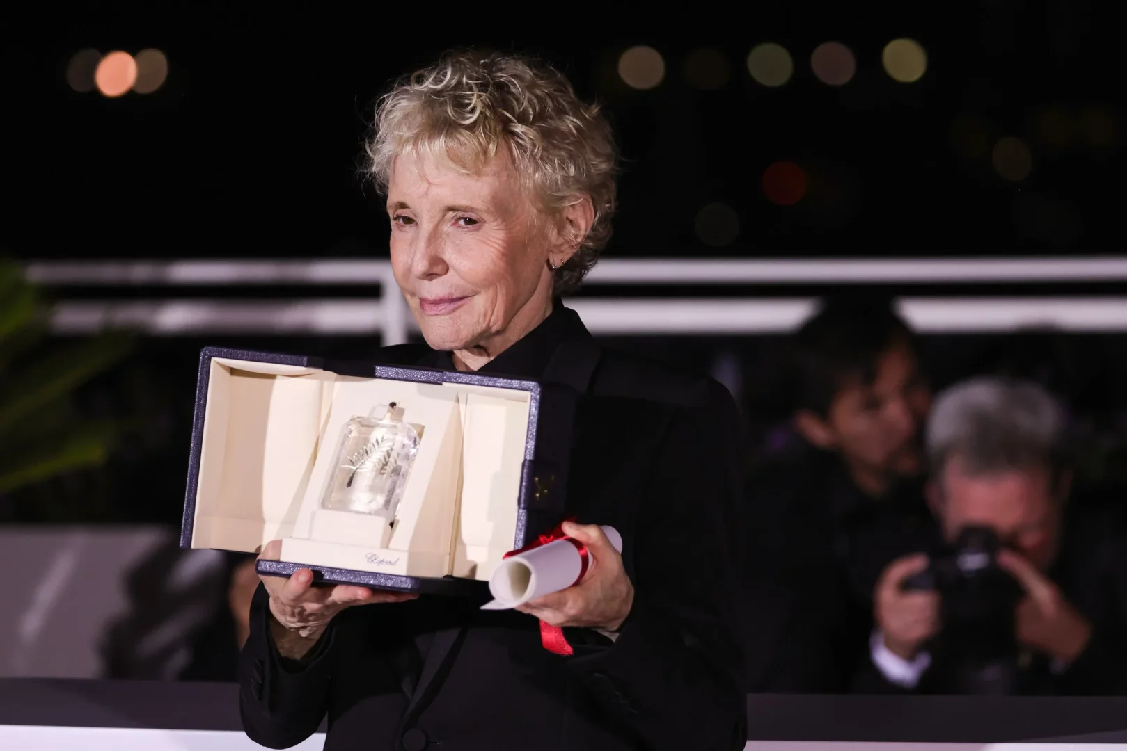 Ünlü film yönetmeni Claire Denis, kıvırcık açık renk saçlarıyla, siyah bir ceket giymiş olarak, lacivert kutusunun krem rengi içindeki Chopard yazılı kristal Palmiye Dalı ödülünü ve kırmızı kurdeleli rulo halindeki sertifikasını gururla sergilerken hafifçe gülümsüyor, karanlık arka planda bulanık ışıklar ve insanlar seçiliyor.