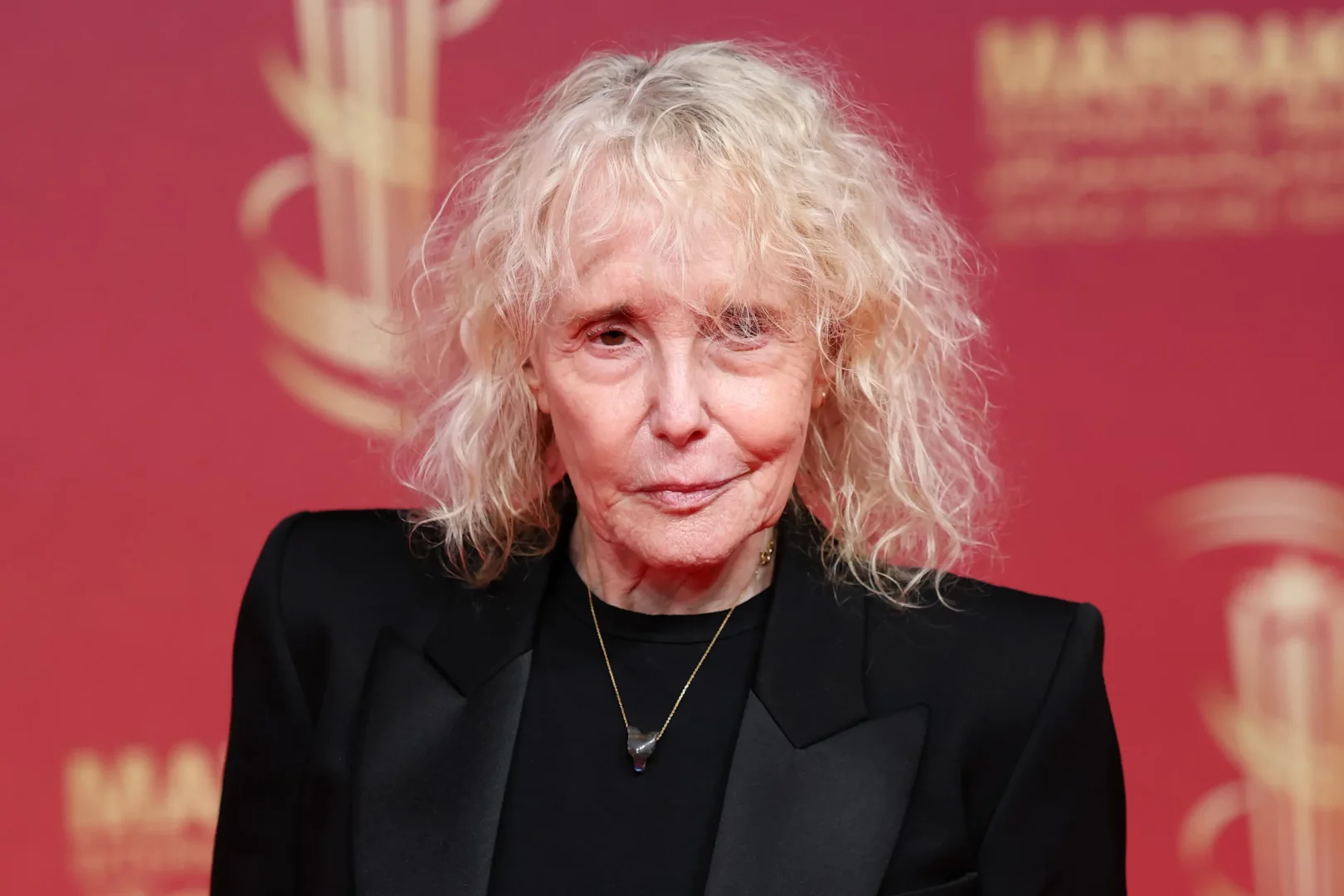 Marrakech Film Festivali veya benzeri bir kırmızı halı etkinliğinde, dağınık sarı saçları, siyah blazer ceketi ve boynunda ince bir kolyeyle objektife hafifçe gülümseyen deneyimli yönetmen Claire Denis'in yakın çekim portresi.