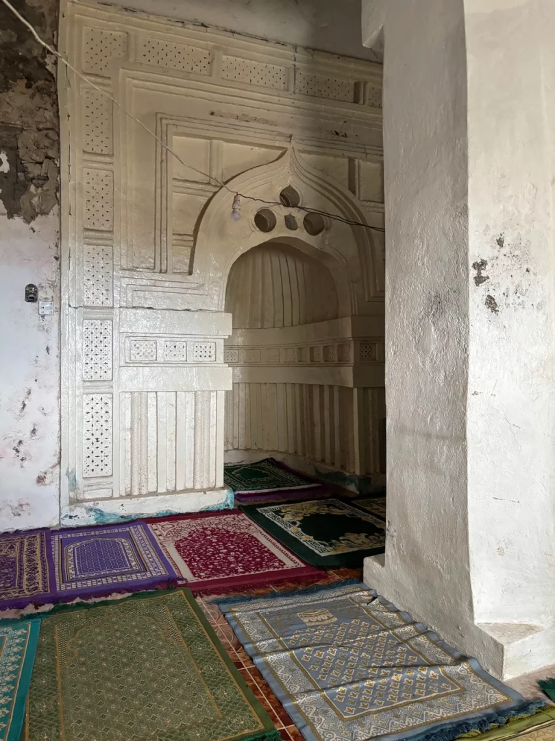 Bu görüntü, UNESCO Dünya Mirası Listesi'ne yeni eklenen, Hint Okyanusu'ndaki Komor Sultanlıkları medinalarının anaerkil İslami gelenekleri ve estetik ada yaşam formlarını yansıtan tarihi dokusundan bir kesit sunuyor; yıpranmış duvarlarına rağmen işlemeli açık renk bir mihrap ve zemine serilmiş renkli seccadeler, mekânın ruhani atmosferini vurguluyor.