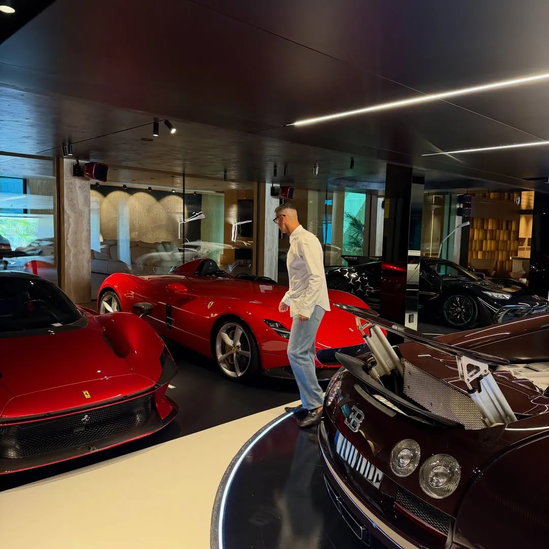 Cristiano Ronaldo, lüks ve modern bir garajda, açık renkli giysileriyle kendi geniş araba koleksiyonundaki parlak kırmızı bir spor arabanın ve diğer lüks modellerin arasında yürürken veya onları incelerken görülüyor.