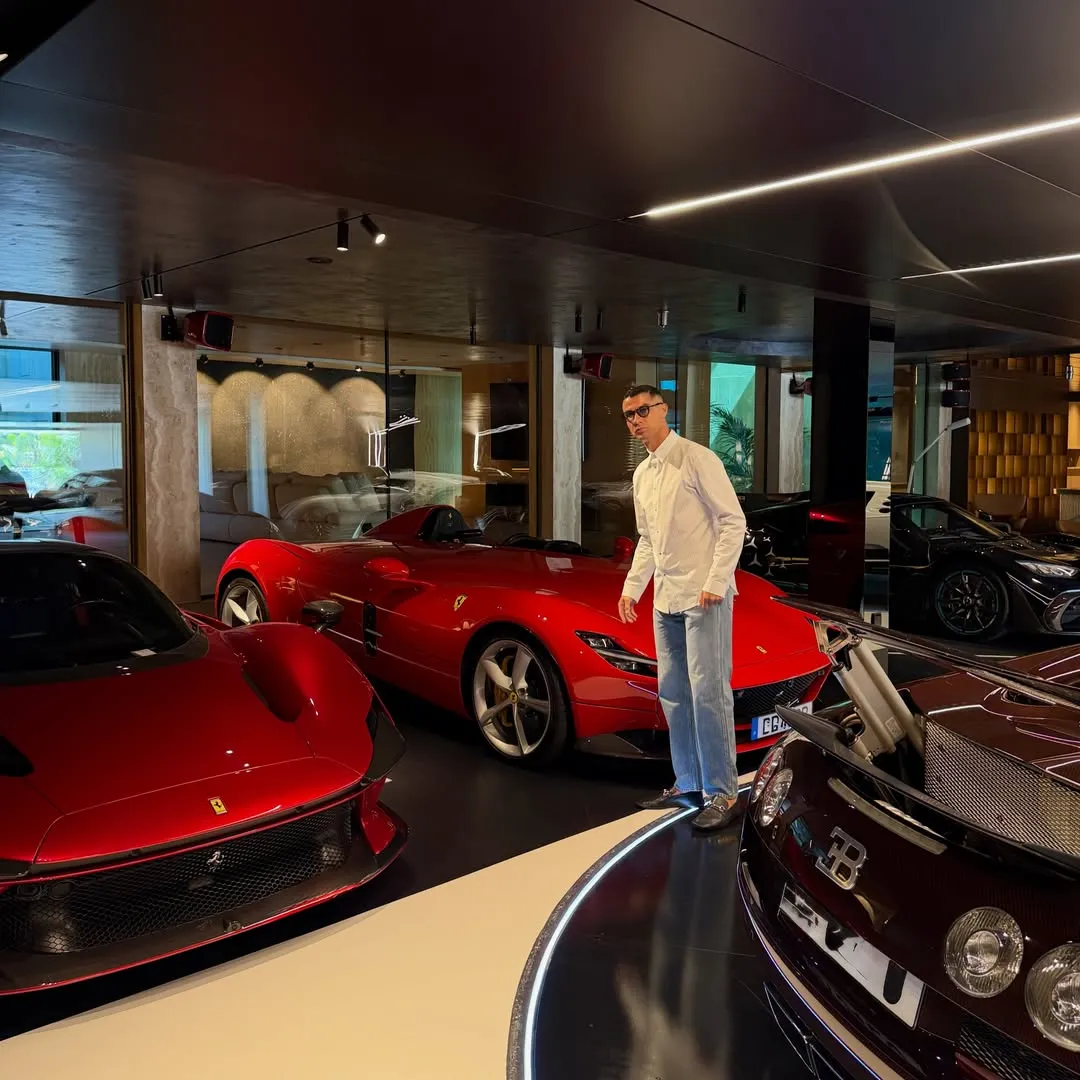 Ünlü futbolcu Cristiano Ronaldo, modern ve şık garajında, parlak kırmızı iki Ferrari ve ön planda bir Bugatti'nin de bulunduğu lüks spor arabaları koleksiyonunun ortasında, beyaz bir gömlek ve açık mavi kot pantolon giymiş şekilde objektife bakarak duruyor.