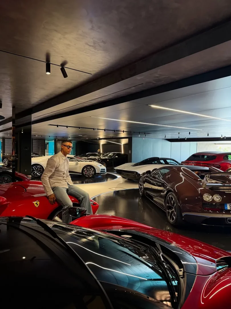 Cristiano Ronaldo, şık ve modern tasarımlı geniş bir garajda, kırmızı bir Ferrari spor arabanın önünde rahat bir şekilde oturmuş, gözlüğünü takmış ve etrafında kendisine ait olduğu anlaşılan farklı marka ve modelde birçok lüks otomobil sergilenmektedir.