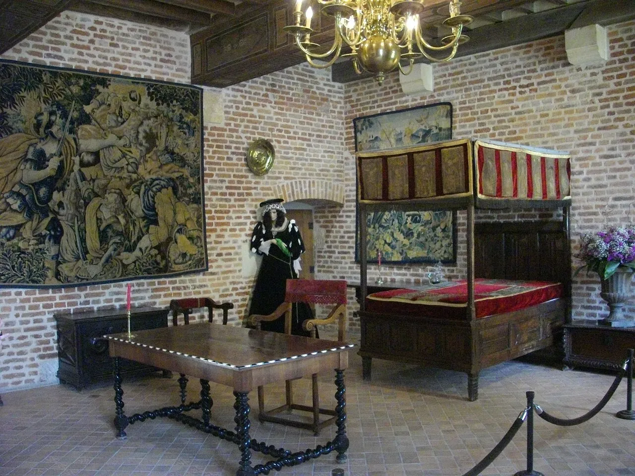 Clos Lucé'deki bir odanın tuğla duvarları arasında, büyük bir duvar halısı, kırmızı ve altın rengi yatak takımlarıyla süslenmiş dört direkli bir yatak, döneme uygun giyimli bir manken ve zarif ahşap mobilyalarla Da Vinci'nin yaşadığı dönemin atmosferini yansıtan zengin bir iç mekan görünümü.