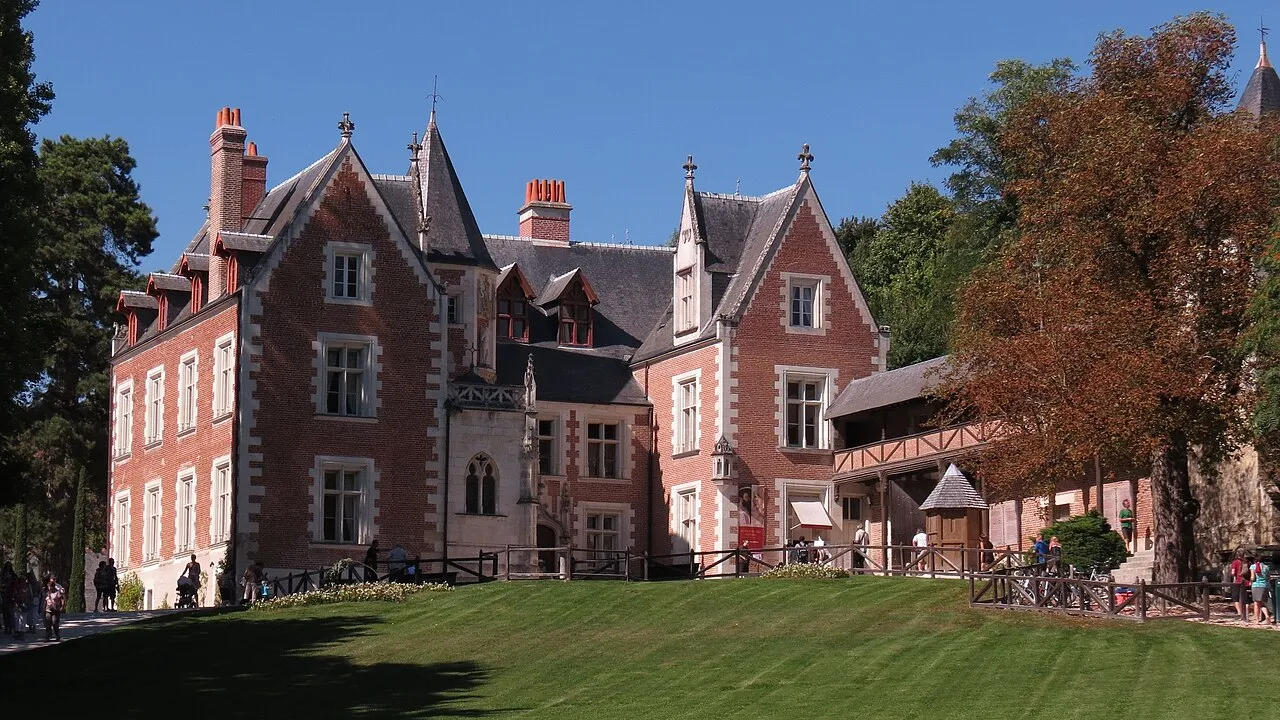 Güneşli bir günde yemyeşil bir çim alana bakan, kırmızı tuğla duvarları ve beyaz taş süslemeleriyle dikkat çeken, sivri çatılı ve kuleli büyük bir tarihi bina olan Clos Lucé, çevresinde yürüyen ziyaretçiler ve gür ağaçlarla huzurlu bir atmosfer sunuyor.