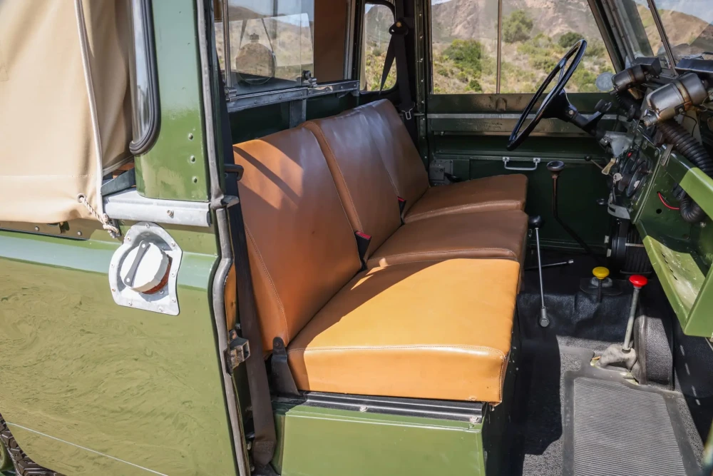 Açık artırmaya sunulacak Daniel Craig'e ait Land Rover Defender Series II arazi aracının taba rengi üçlü ön koltuklarını, direksiyon simidini ve vites kollarını öne çıkaran bu iç mekan tasarımı görüntüsünde, aracın yeşil dış rengi, bej tente tavanı ve arka plandaki tepelik arazi manzarası dikkat çekmektedir.