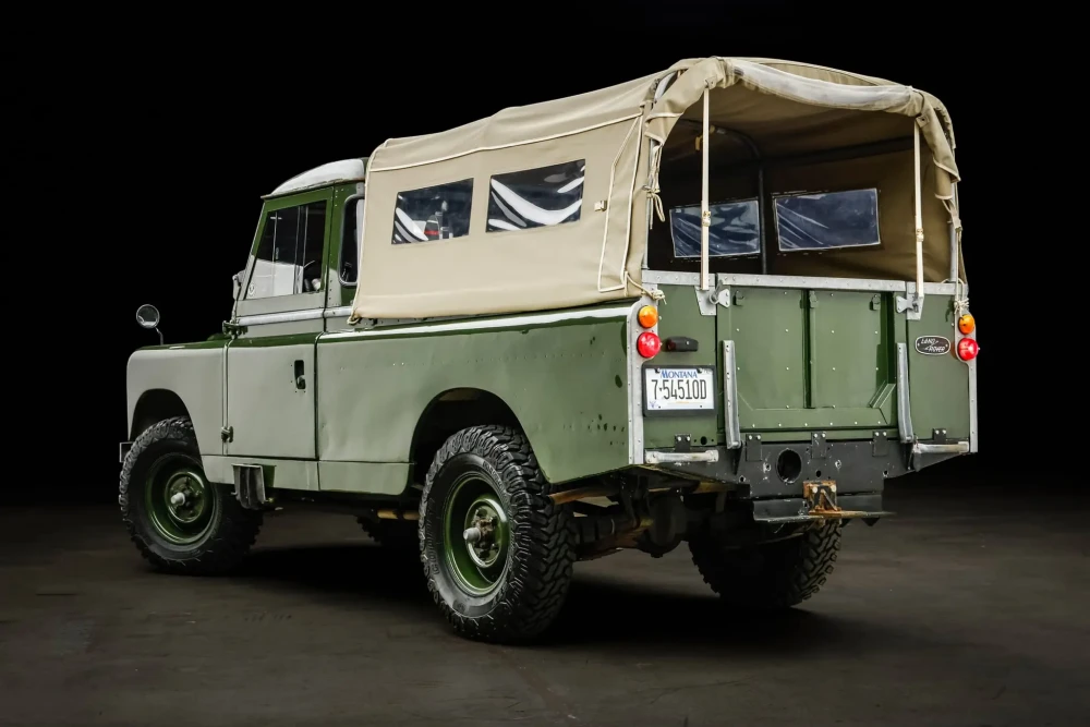 Daniel Craig'in Land Rover Defender Series 2 modelinin arka 3/4 görünümünü sunan bu fotoğrafta, khaki yeşili gövdesi ve bej renkli branda tavanı ile araç, klasik arazi tekerlekleri ve Montana plakasıyla karanlık bir stüdyo ortamında açık artırma için dikkat çekici bir şekilde sergileniyor.