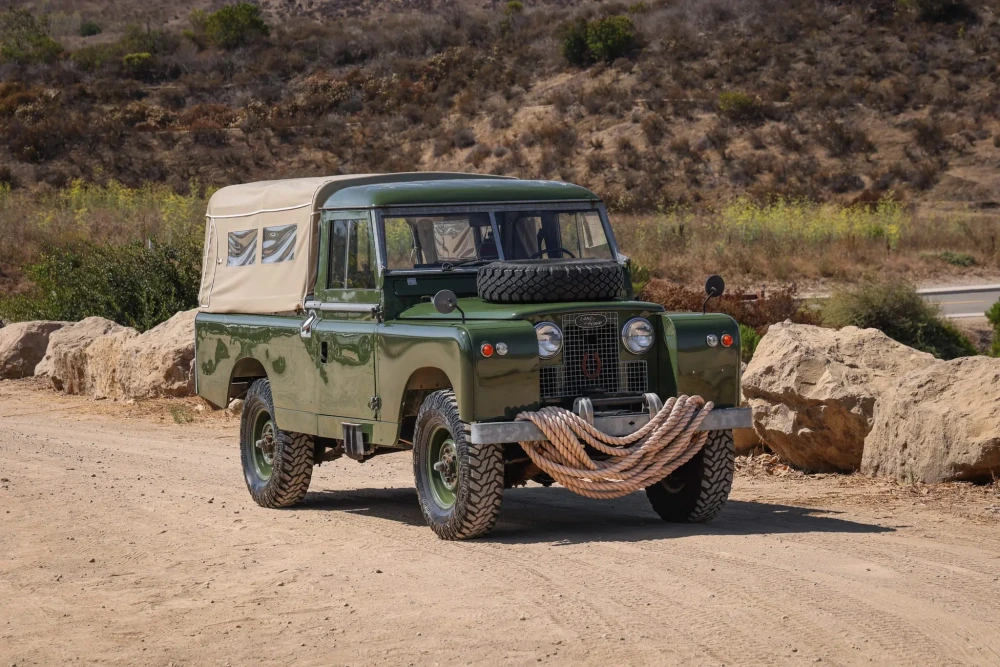 Yeşil renkli, Daniel Craig ile ilişkilendirilen Land Rover Defender Series 2 arazi aracı, kahverengi tepelerin ve büyük kayaların çevrelediği toprak bir yolda duruyor; bej kanvas tavanı, kaputundaki yedek lastiği ve ön tamponuna sarılı kalın halatıyla karakteristik dış tasarımını açıkça sergiliyor ve açık artırma için hazır görünüyor.