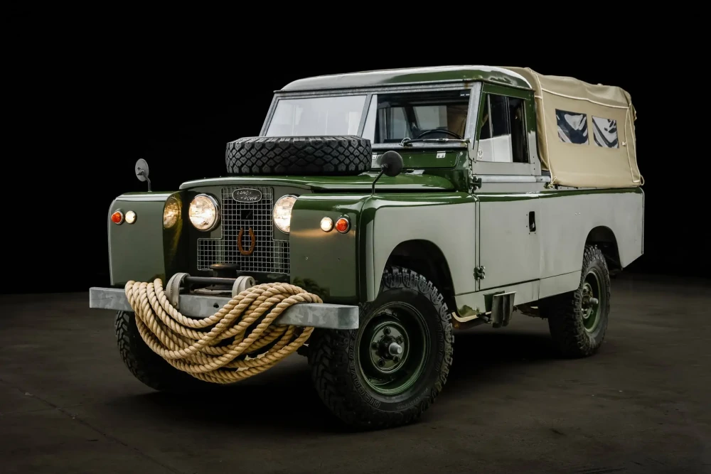 Zeytin yeşili ve açık renk kanvas tente detaylarına sahip, kaputunda yedek lastik taşıyan ve ön tamponunda kalın bir halatın estetik bir şekilde sarılı olduğu, Daniel Craig'in Land Rover Defender Serisi 2'sinin ikonik dış tasarımını vurgulayan, koyu bir zemine karşı etkileyici bir şekilde sergilenen klasik arazi aracı.