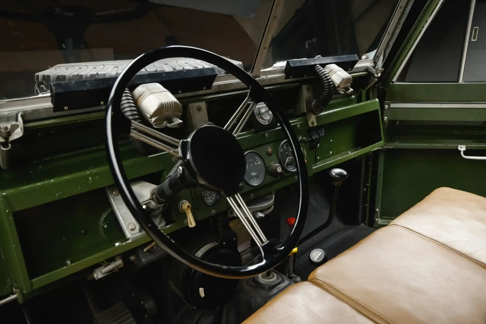 Daniel Craig'in Land Rover Defender Series 2si aracının iç mekanını gösteren bu detaylı fotoğrafta, ortada yer alan siyah direksiyon simidi, askeri yeşil renkteki sade ön konsol, üzerinde klasik göstergeler ve sağda kahverengi benç koltuk göze çarparken, aracın zamansız ve sağlam tasarımını vurgulayan unsurlar sergileniyor.