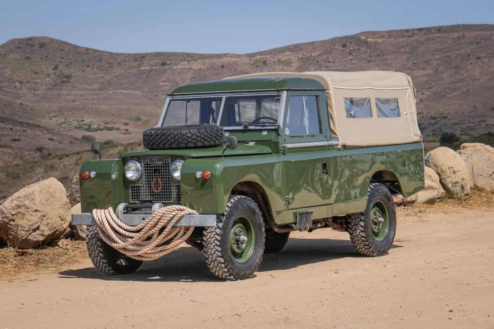 Kurak bir tepelik arazideki toprak yolda, bej renkli tentesi, kaputundaki yedek lastiği ve ön tamponuna sarılı halatıyla off-road kullanımına hazır bekleyen bu klasik zeytin yeşili Land Rover araç, yaklaşan açık artırma etkinliğinin odak noktası.