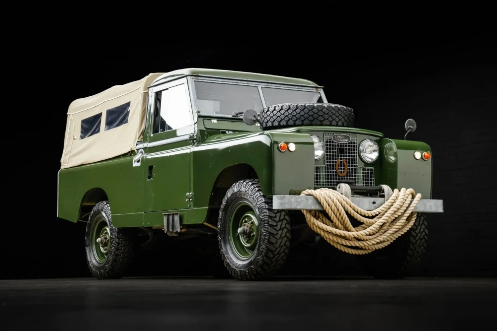 Daniel Craig'in Land Rover Defender Series II modeli, koyu yeşil gövdesi, bej kumaş tentesi, kaputundaki yedek lastiği ve ön tamponuna sarılı kalın halatıyla açık artırma öncesi çekilmiş dikkat çekici bir fotoğraf.