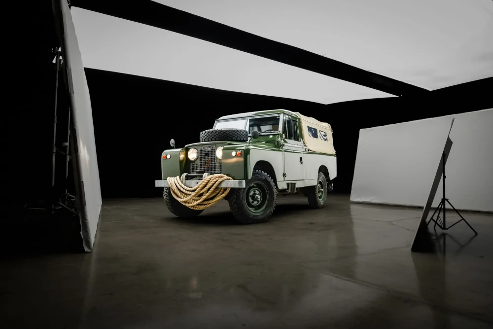 Stüdyo ortamında, ön tamponuna sarılmış kalın bir halat, kaputunda yedek lastiği ve bej tente çatısıyla, yeşil beyaz renkli, Daniel Craig'in Land Rover Defender Series 2 açık artırma aracı sergileniyor.