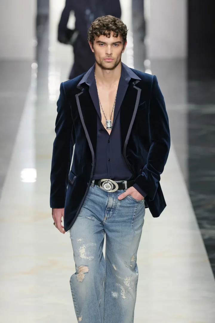Podyumda yürüyen, koyu mavi kadife Dolce & Gabbana blazer ceketi, içindeki lacivert gömleği, yırtık açık mavi kot pantolonu, büyük metal tokalı kemeri ve uzun kolyesiyle şık ve modern bir tarz sergileyen genç erkek model.