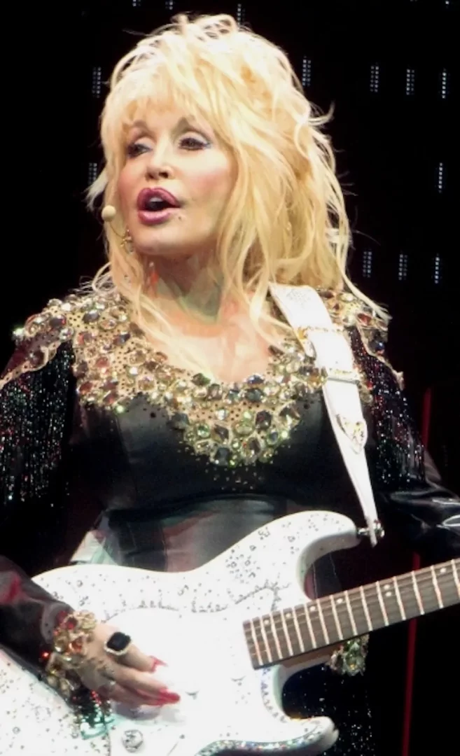 Dolly Parton, 2014 yılında Köln Lanxess Arena'daki konserinden bir karede, sarı dalgalı saçları, ışıltılı işlemeli siyah sahne kıyafeti ve beyaz elektro gitarıyla coşkulu bir performans sergiliyor.